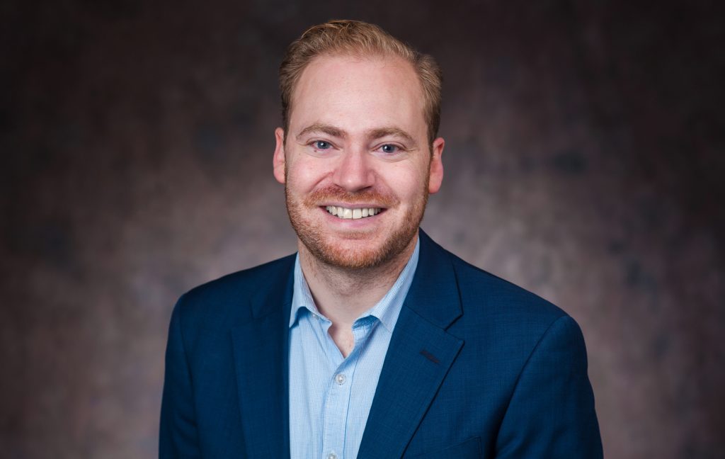 Photo of Andy MacCracken smiling in a button-up shirt and blazer.