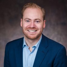 Photo of Andy MacCracken smiling in a button-up shirt and blazer.